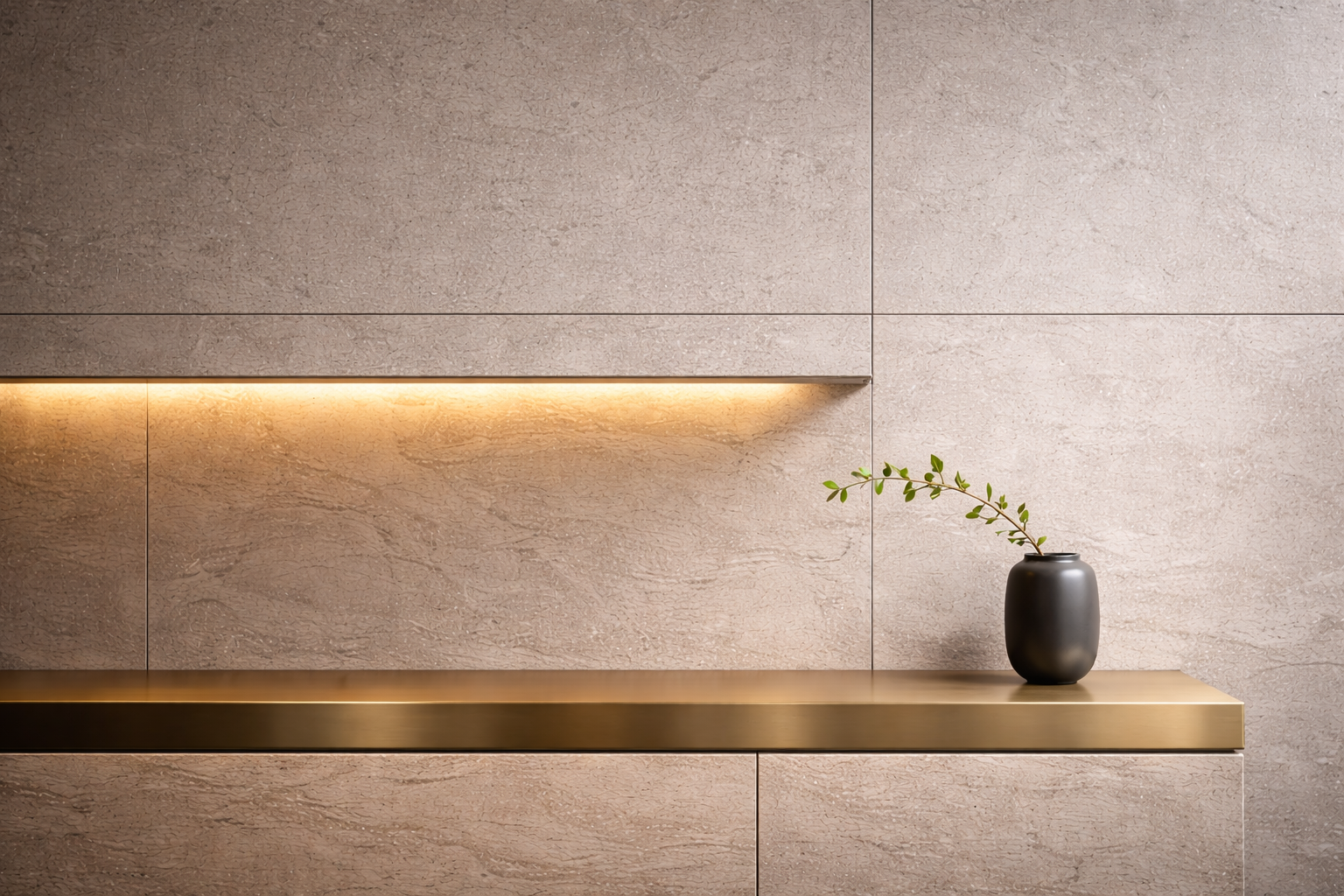 Mensola in metallo dorato con vaso nero contenente ramoscello con foglie verdi su sfondo di parete beige con illuminazione calda.