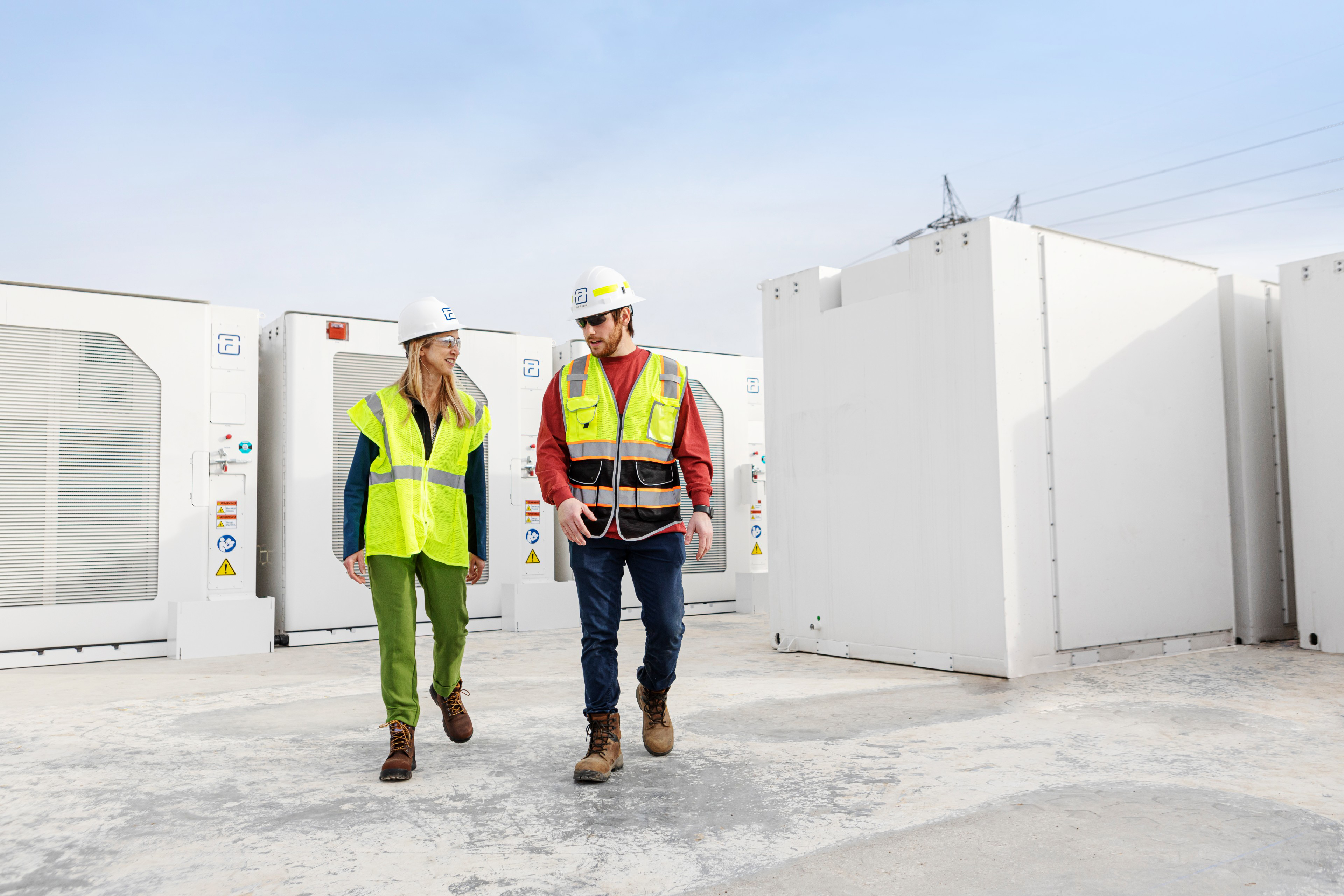 Two construction workers wearing hard hats and high-visibility vests walking and talking at an industrial site with large white equipment units in the background.