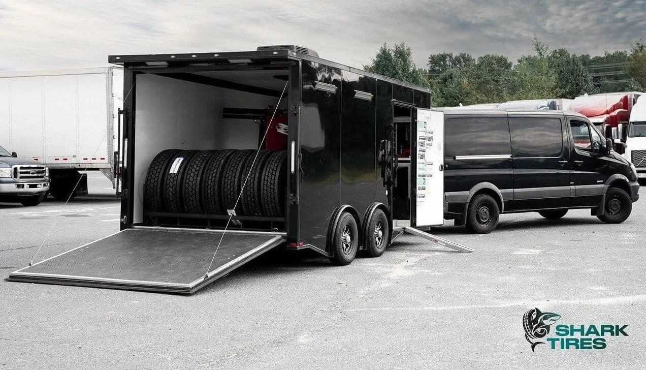 A black tire trailer with an open ramp, showcasing stacked tires inside, parked beside a delivery van.