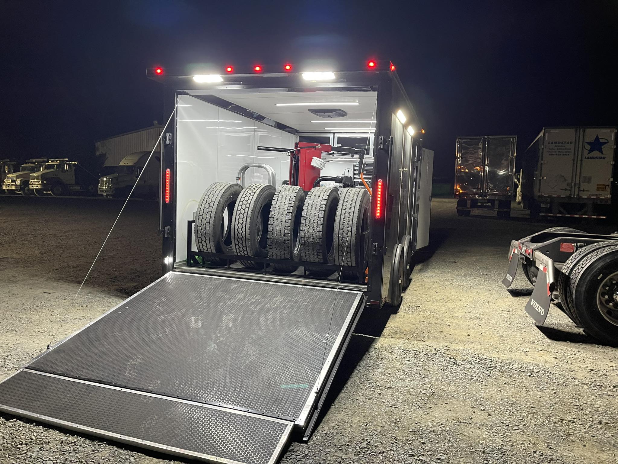 A dimly lit trailer opens to reveal stacked tires, ready for loading. Nearby trucks are parked in the night.