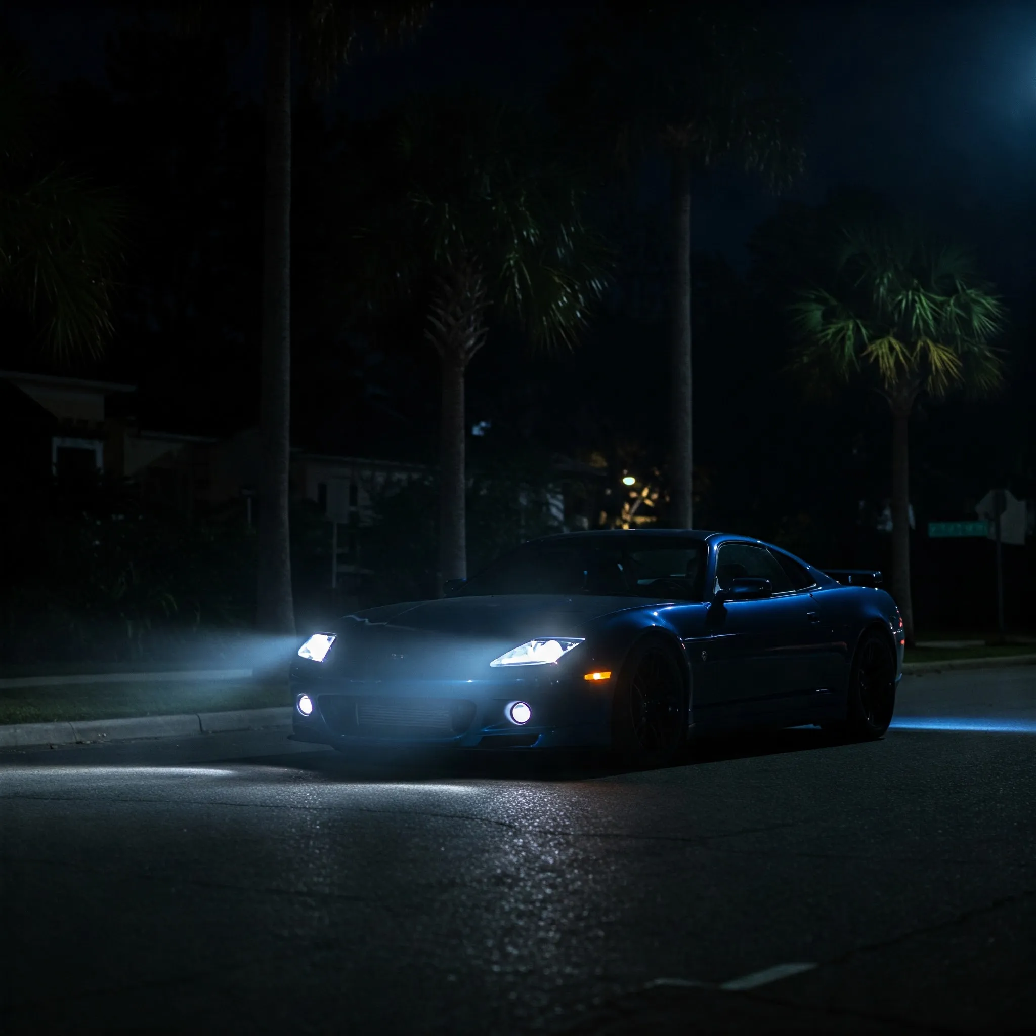 A sleek blue car parked on a dimly lit street at night, illuminated by its bright headlights. Palm trees stand nearby.