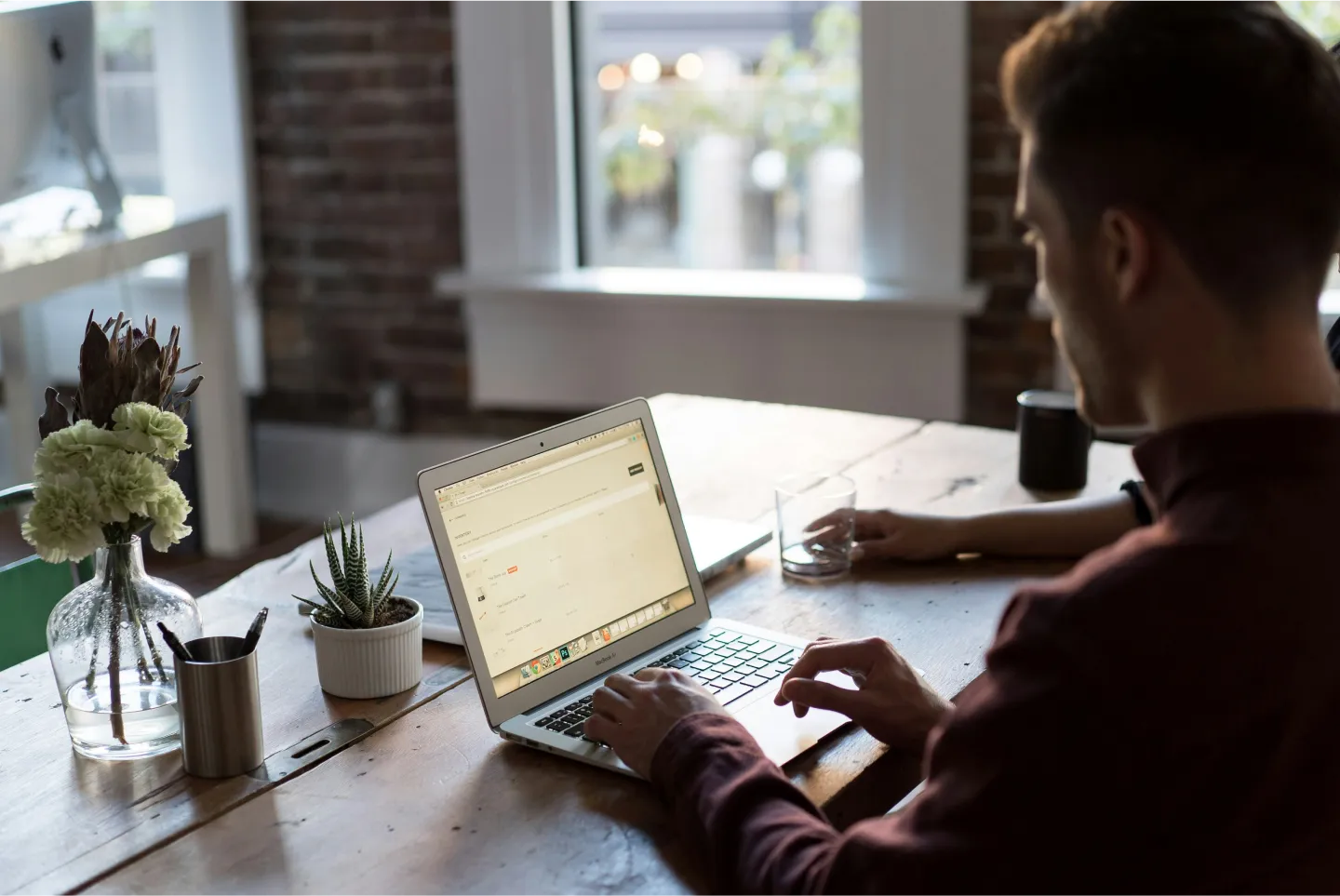 A person working on a laptop at a wooden table, surrounded by plants and a cup of coffee. Sunlight streams through the window.