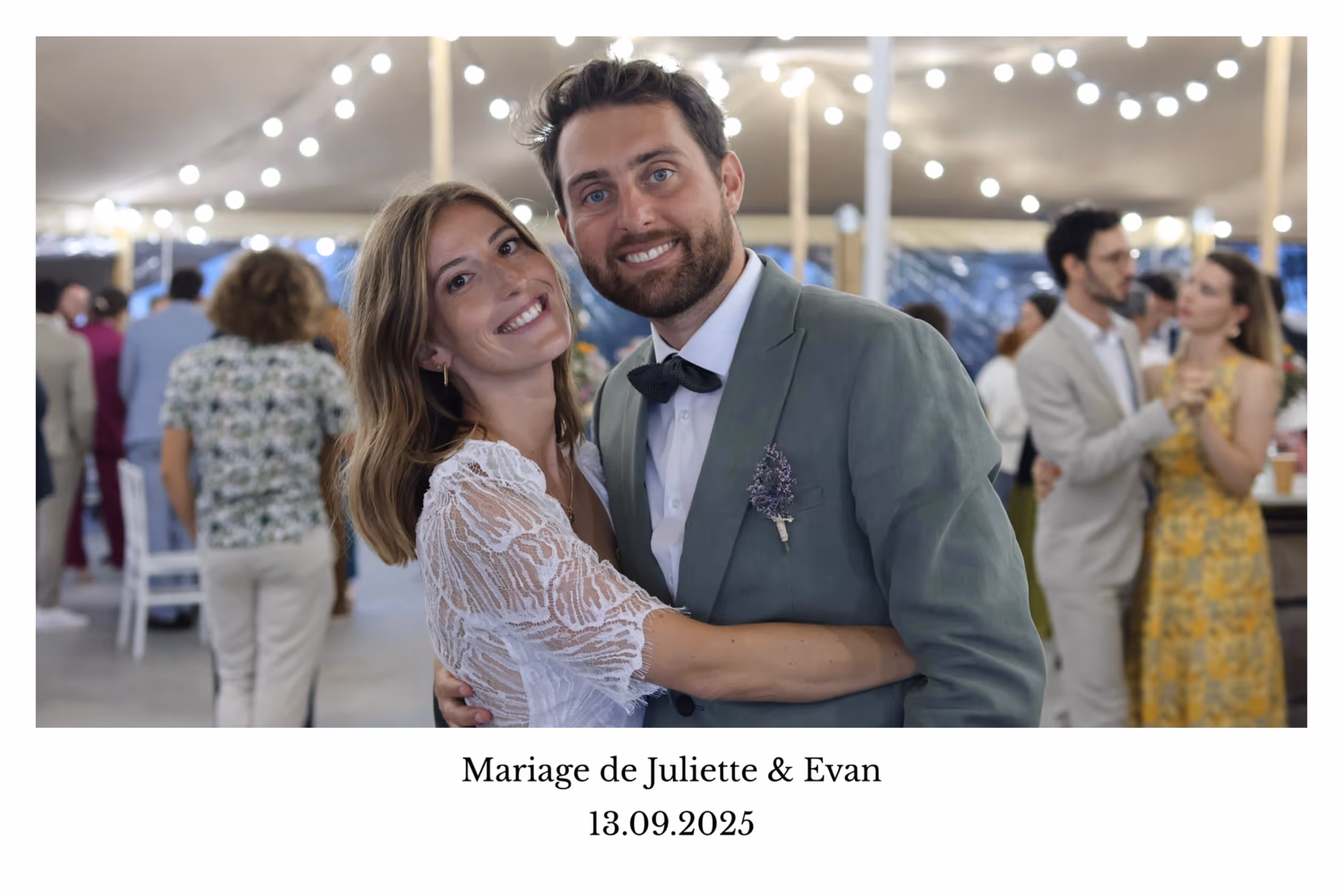 Couple souriant en tenue de mariage s'enlaçant lors de la réception avec des invités dansant en arrière-plan sous des guirlandes lumineuses.