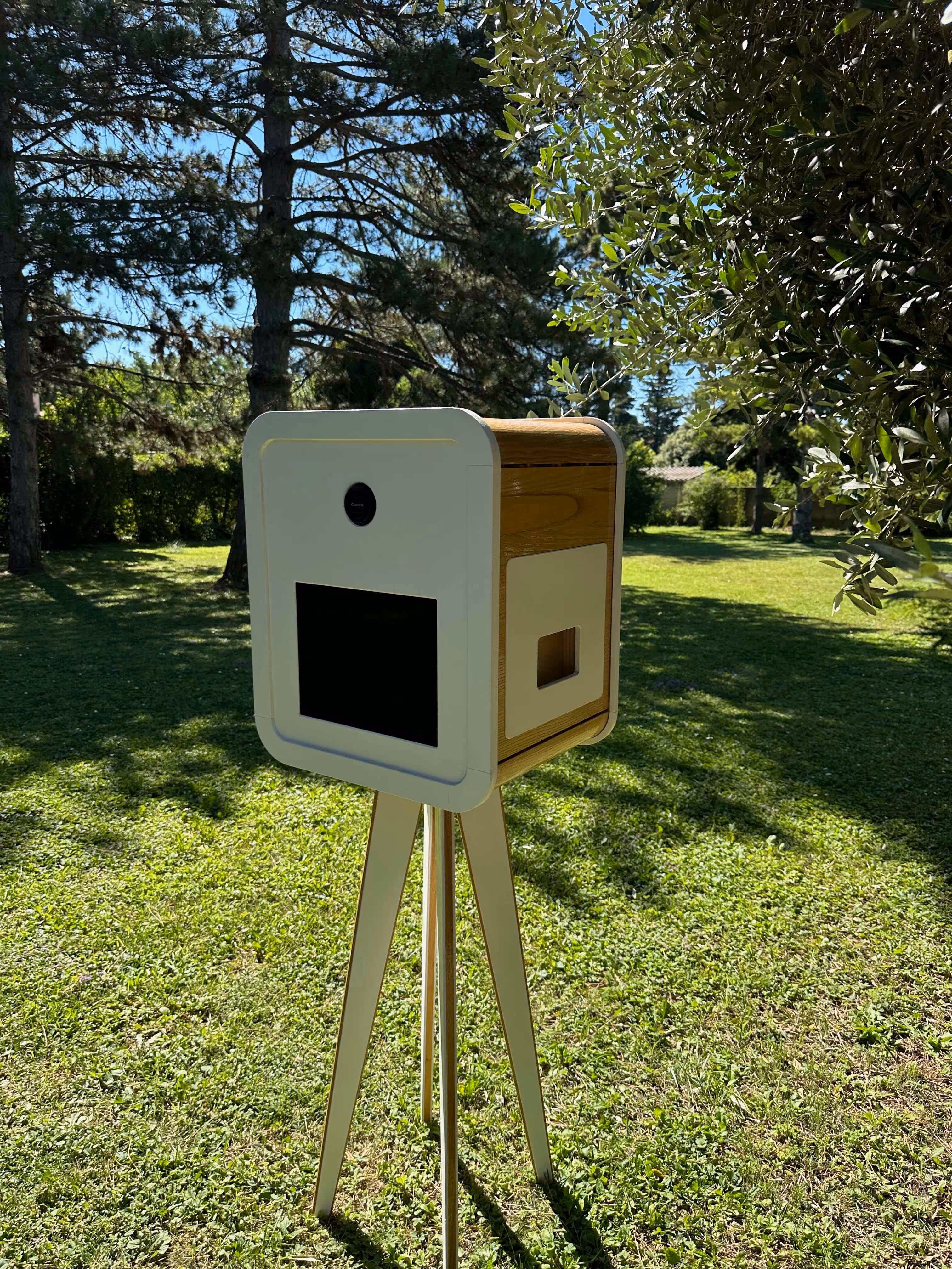 Cabine photo moderne sur trépied blanc et bois installée dans un jardin ensoleillé.