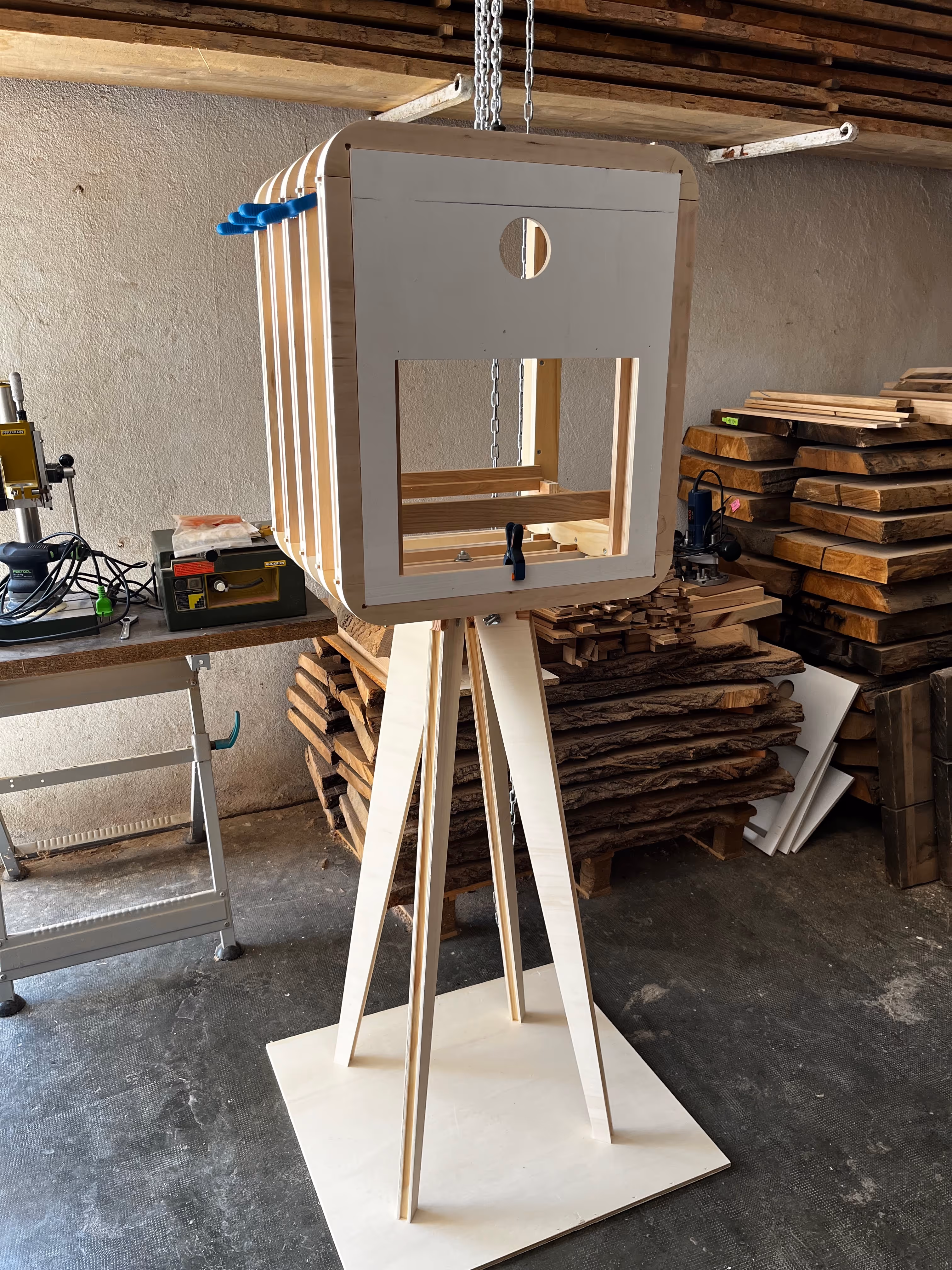 Prototype d'une structure en bois claire sur pied trépied dans un atelier de menuiserie avec des outils et des planches en arrière-plan.