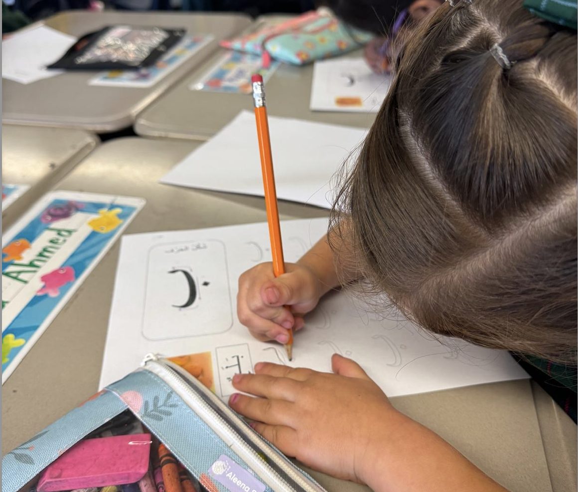 Child holding a pencil and practicing Arabic letters on a worksheet at a classroom desk.
