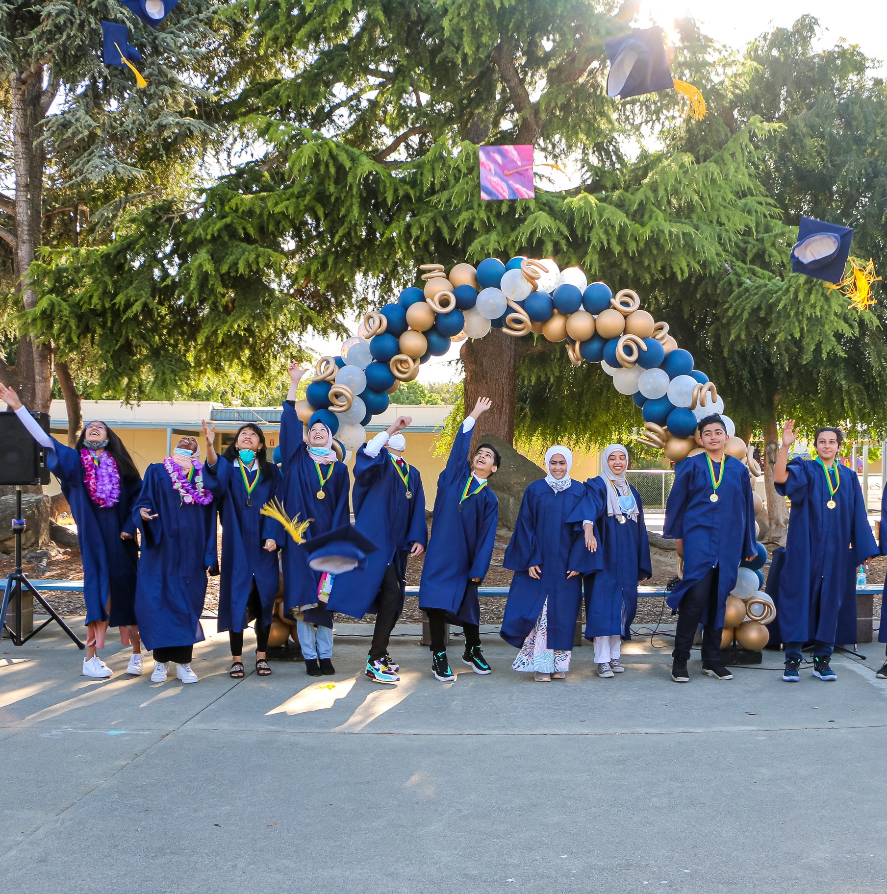 Group of graduates in blue gowns and medals throwing their caps in the air under a blue and gold balloon arch outdoors.