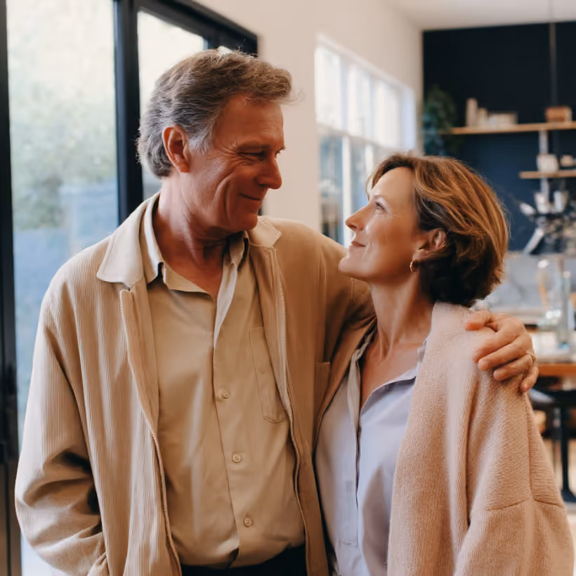 Middle-aged couple smiling and looking at each other warmly in a cozy home setting.