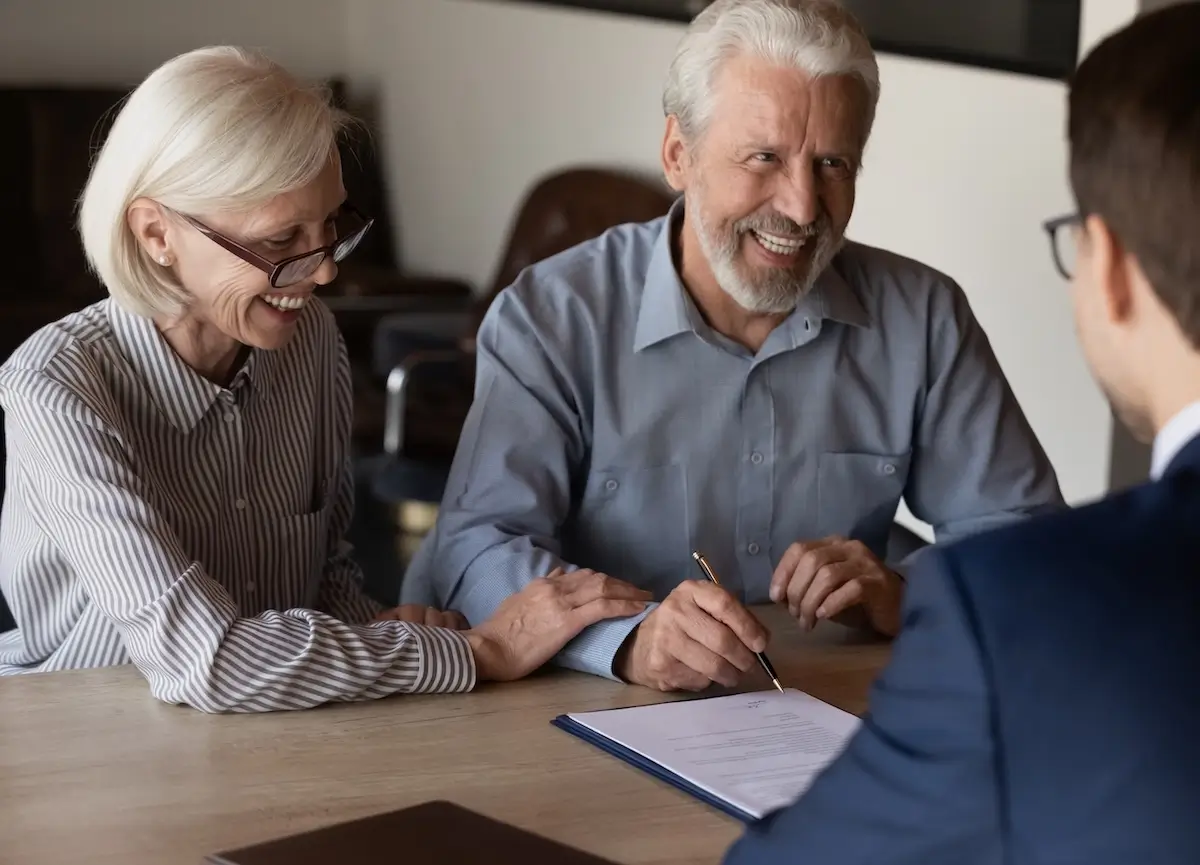 A pleasant elderly couple laughs as they sign a document with their attorney