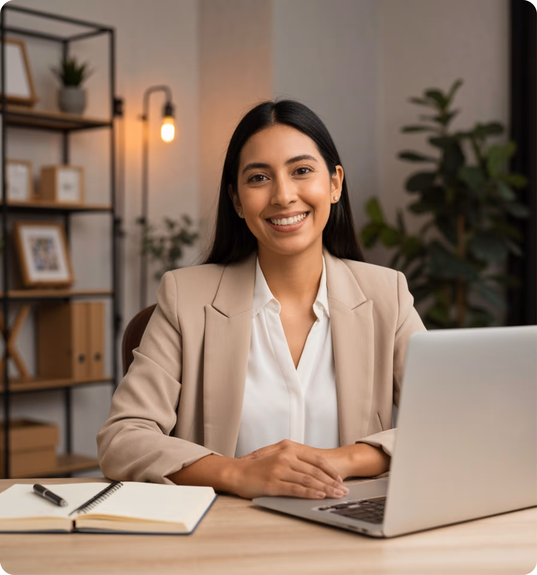 Smiling young businesswoman in beige blazer sitting at desk with laptop and open notebook in modern office.