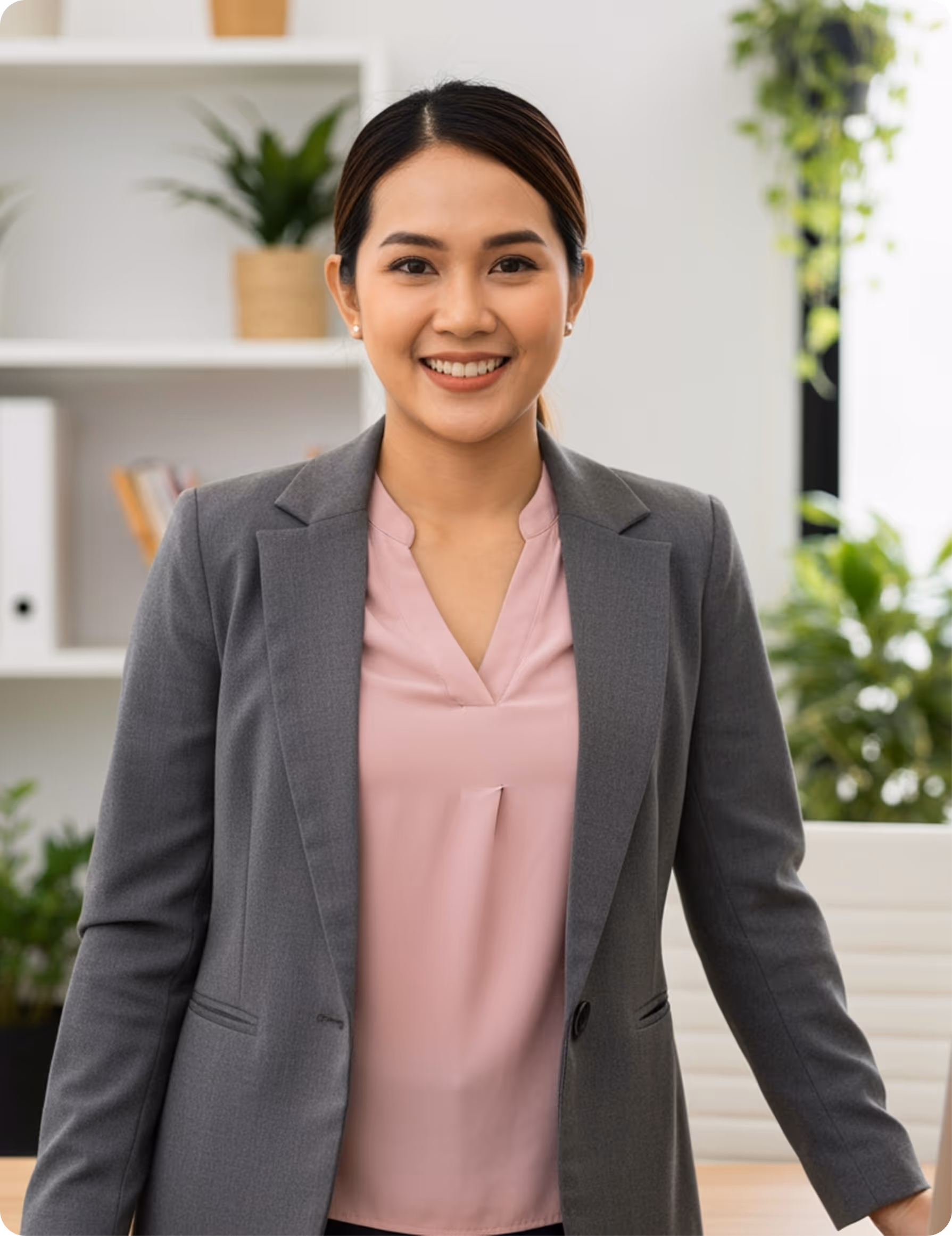 Smiling woman in a gray blazer and pink blouse standing in an office with plants and shelves in the background.