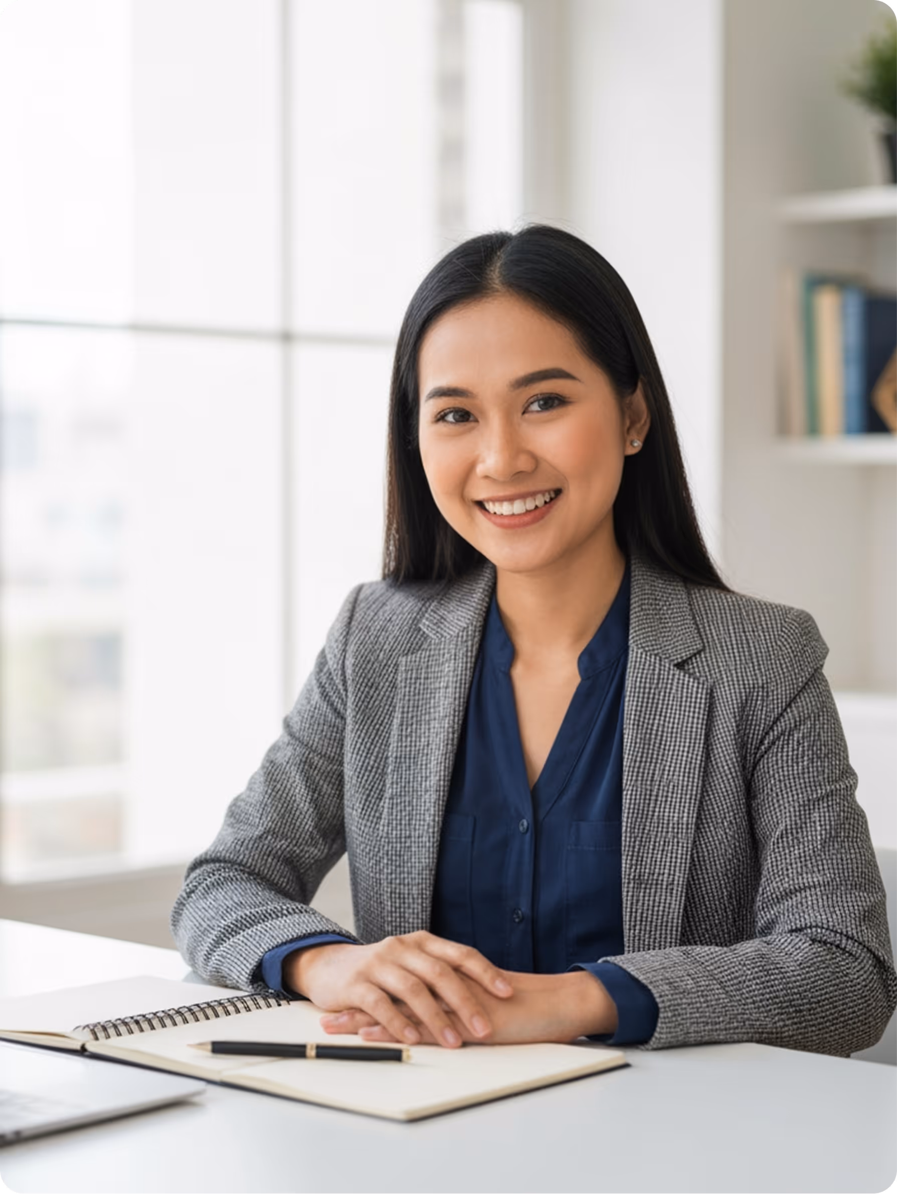 Smiling woman in a gray blazer sitting at a desk with an open notebook and pen.