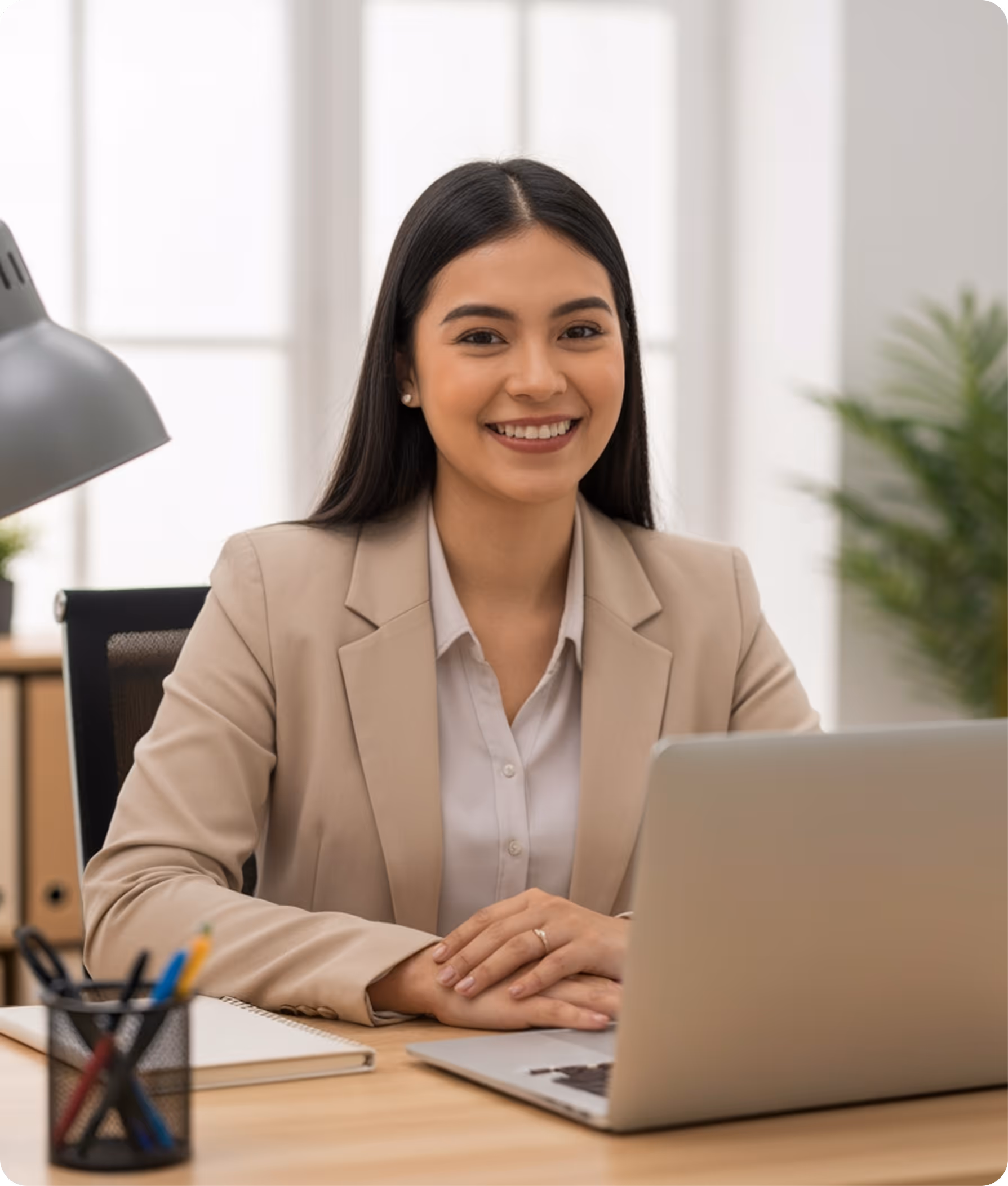 Smiling woman in a beige blazer sitting at a desk with a laptop and stationery in a bright office.