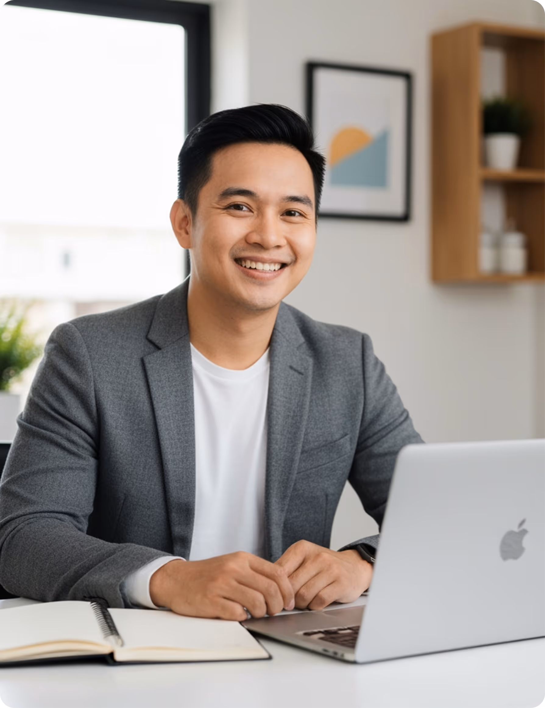 Smiling man in gray blazer sitting at desk with open notebook and Apple laptop.
