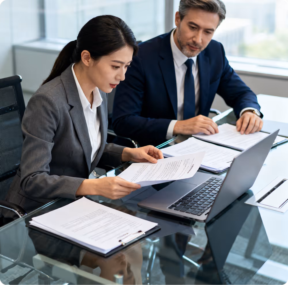 Two business professionals reviewing documents together at a glass conference table with a laptop.