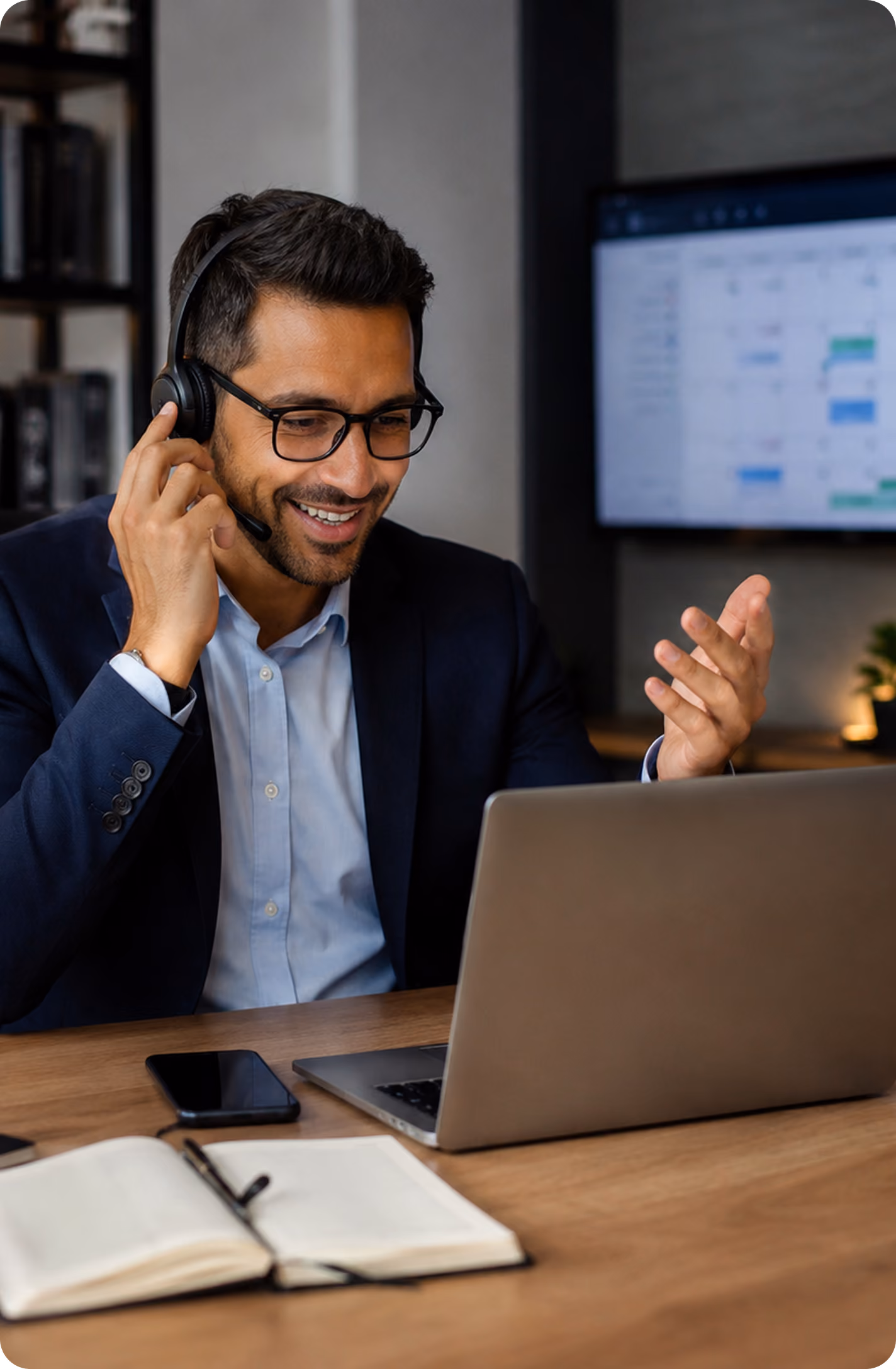 Smiling man wearing glasses and a headset, speaking during a video call on his laptop at a desk.