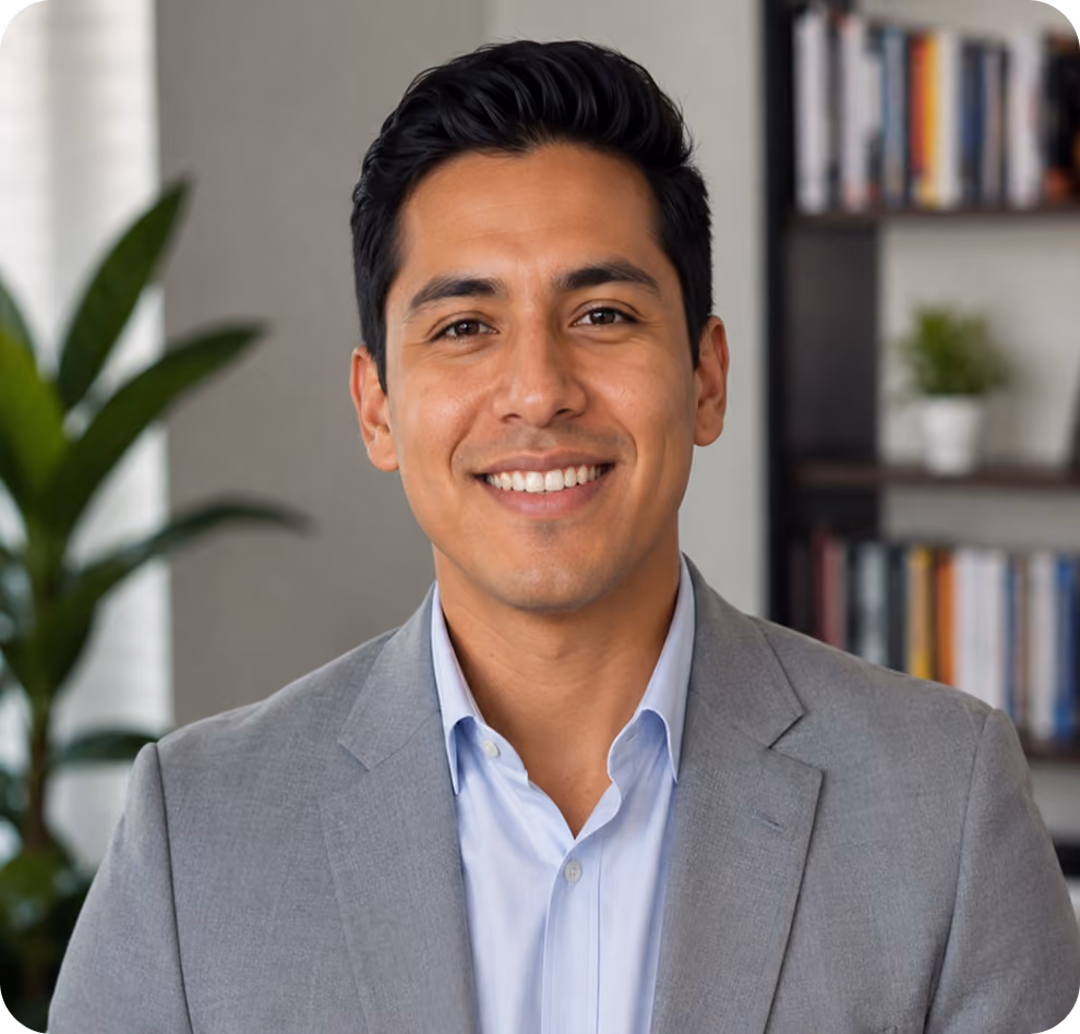 Smiling man in a light gray suit and light blue shirt standing in front of a plant and bookshelf.