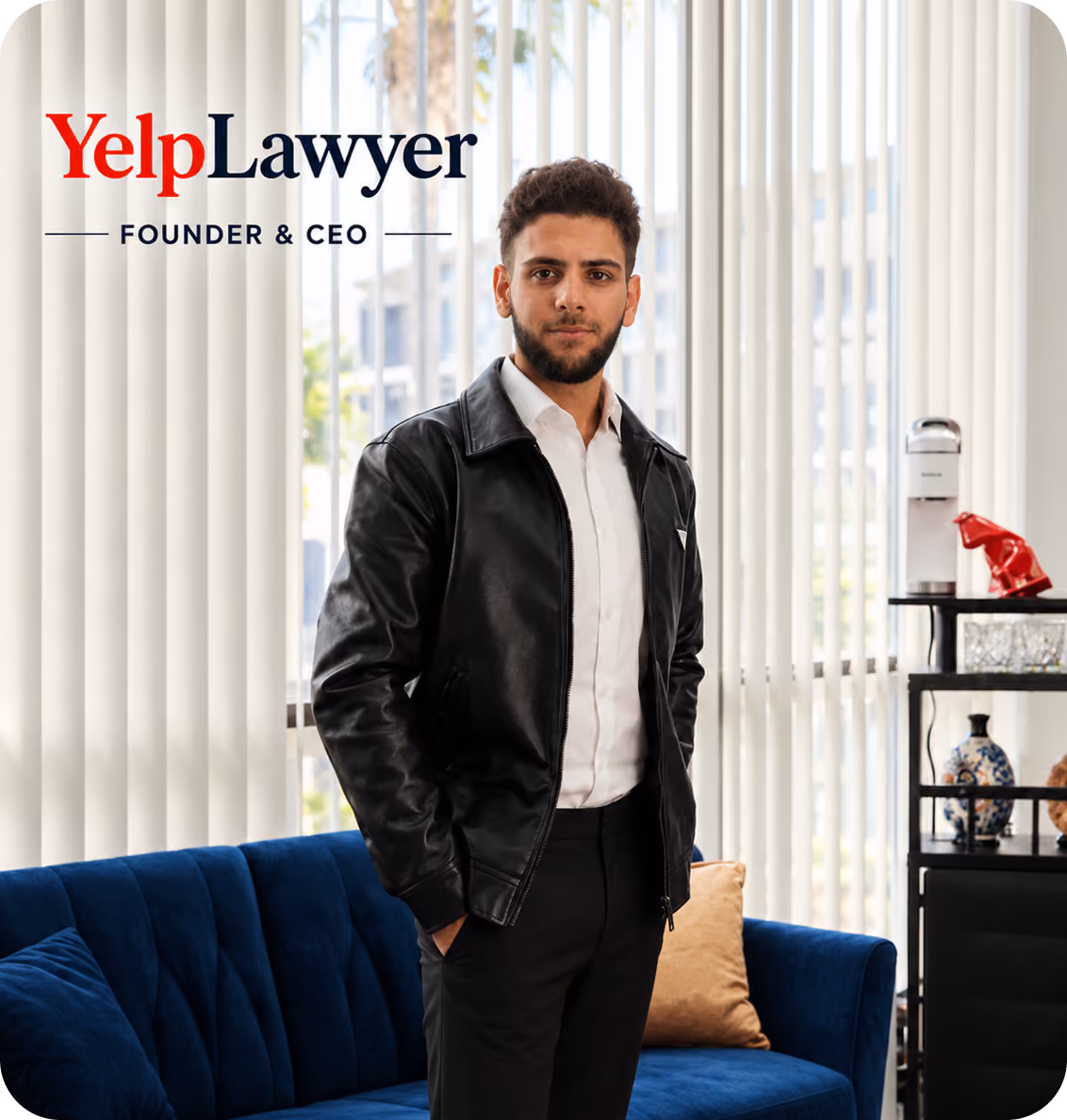 Young man with dark hair and beard wearing a black leather jacket and white shirt, standing with hands in pockets in a modern room with large vertical blinds, a blue couch, and shelves with decorative items.