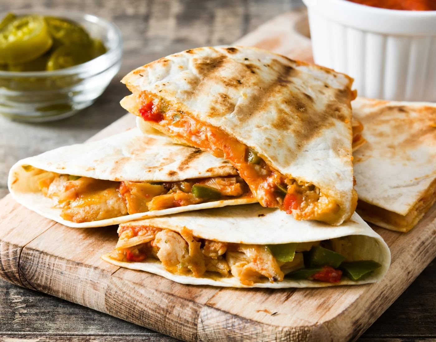 Close-up of grilled chicken quesadilla slices filled with cheese and vegetables on a wooden cutting board, with jalapeños and salsa in the background