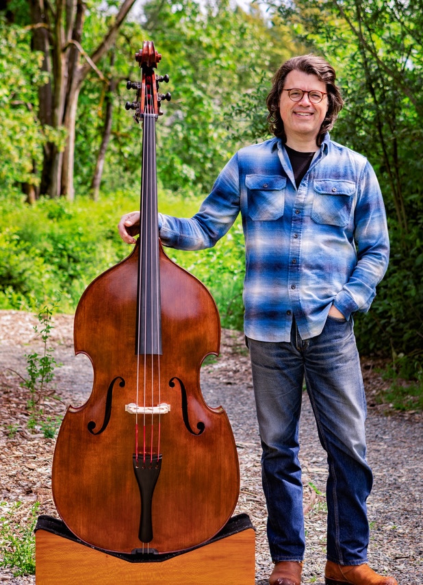 A hand carved double bass made with reclaim wood in a lush green park. Luthier John Koehler stands next to the bass, smiling. 
