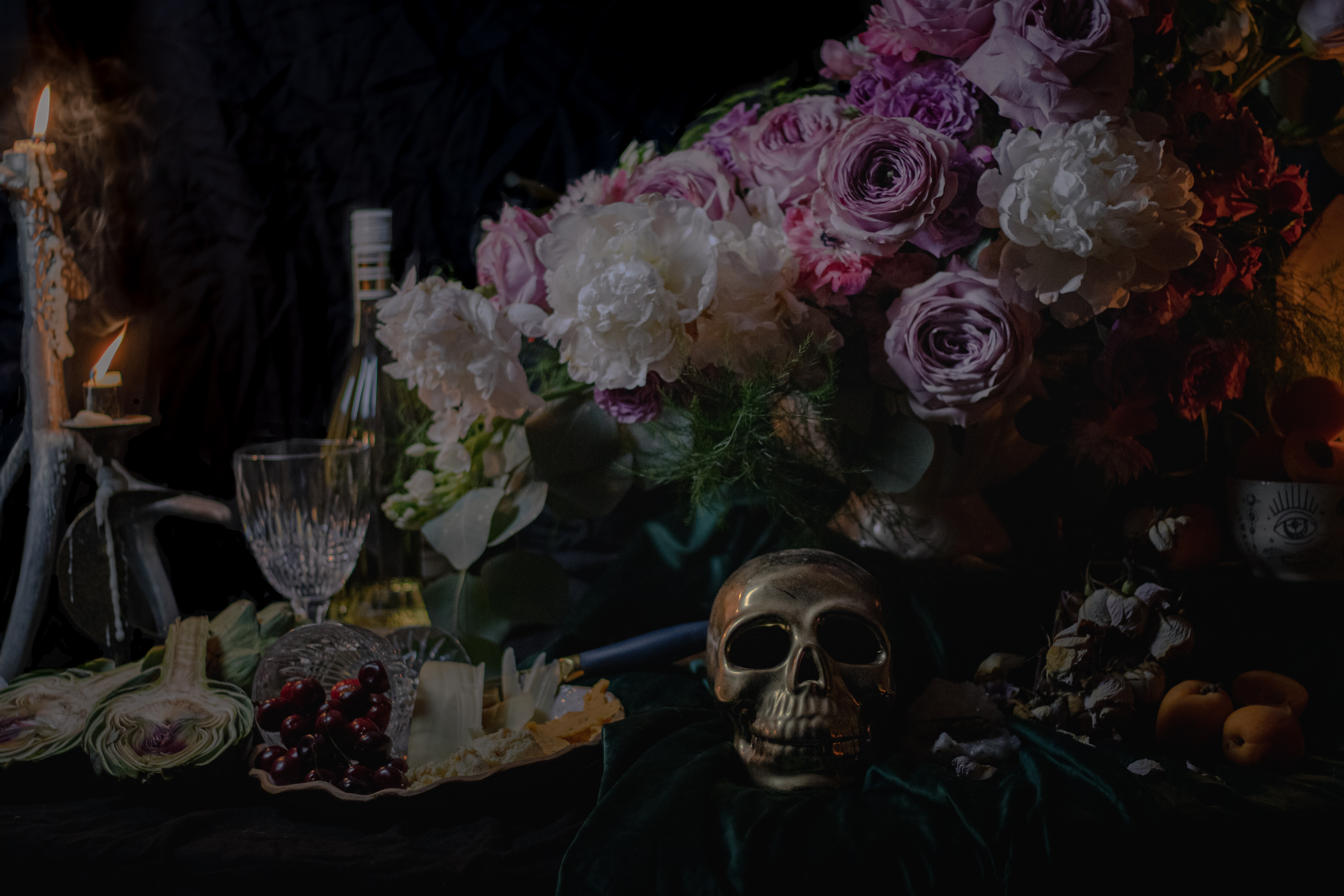 Still life with white and purple flowers, a metallic skull, candles, artichokes, cherries, cheese, a wine glass, and a bottle on a dark table.