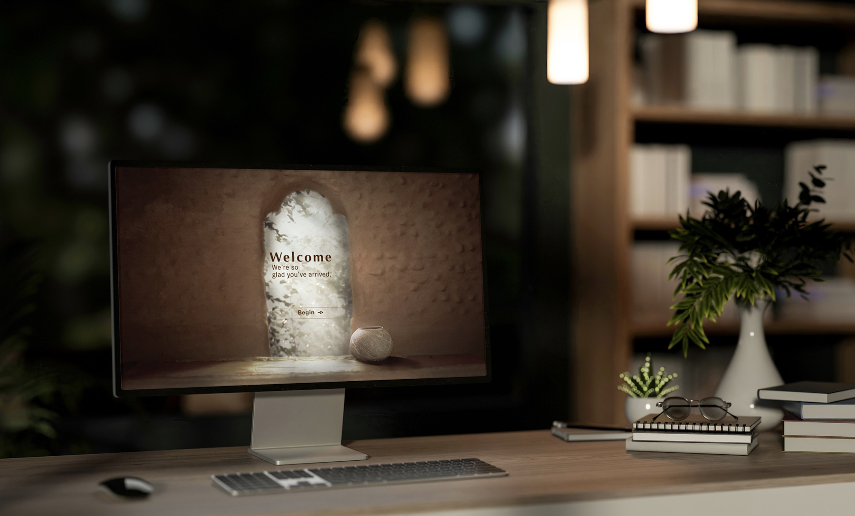 Desk setup with a computer monitor displaying a welcome message, keyboard, mouse, books, glasses, and plants with a blurred bookshelf in the background.