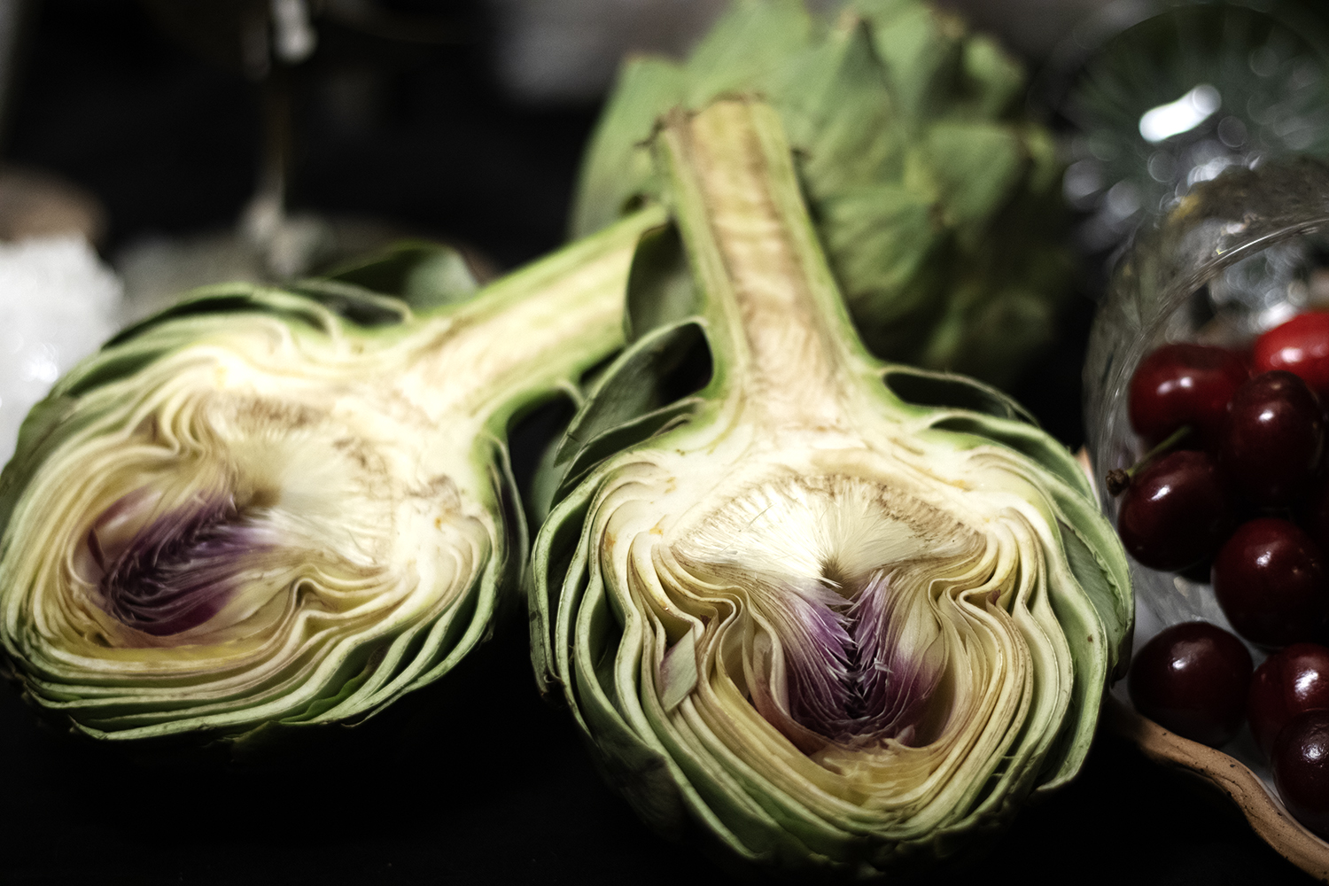 Two halves of a fresh artichoke with purple centers on a dark surface next to a glass bowl of red cherries.