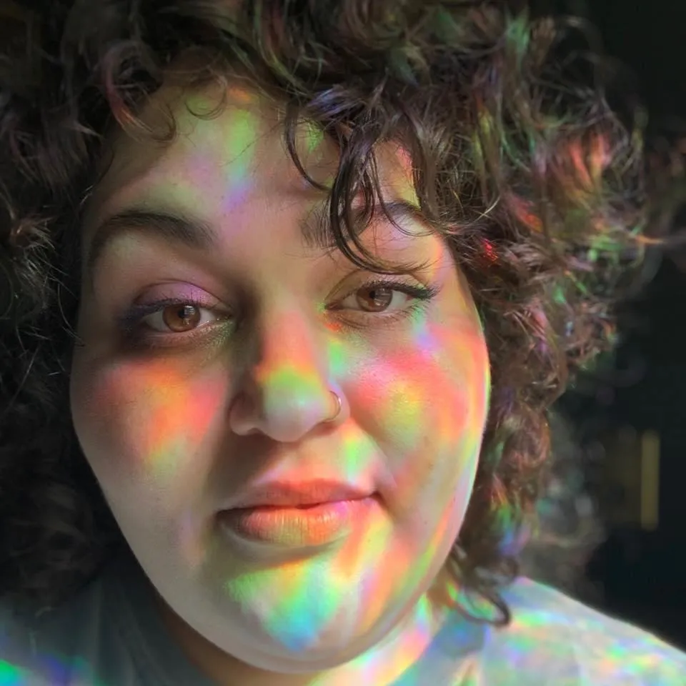 Close-up of a person with curly hair and brown eyes illuminated by colorful rainbow light reflections on their face.