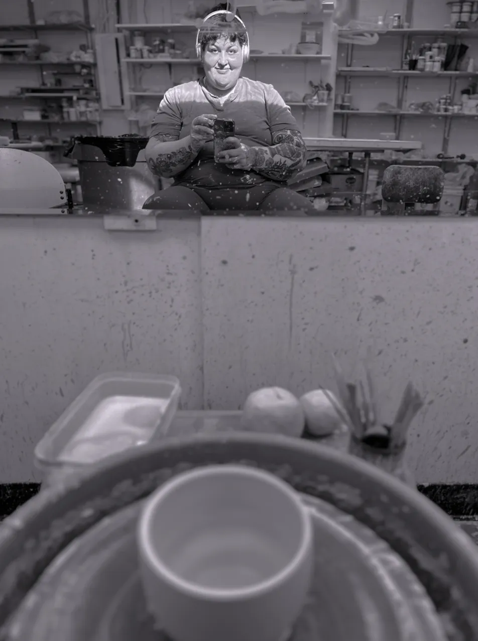 Person with headphones and tattoos taking a selfie in a pottery studio mirror with a pottery wheel and tools in foreground.