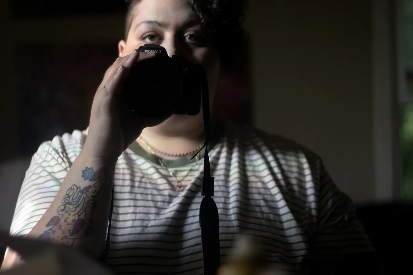 Person taking a mirror selfie with a DSLR camera, wearing a striped shirt and showing a tattoo on the forearm.