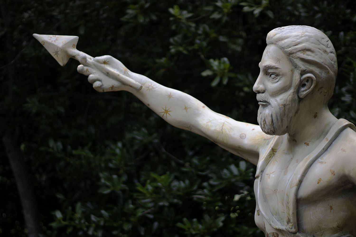White statue of Vulcan, the god of the forge, with an arm extended, holding an arrow, adorned with small star and insect illustrations. Taken at Birmingham Botanical Gardens.