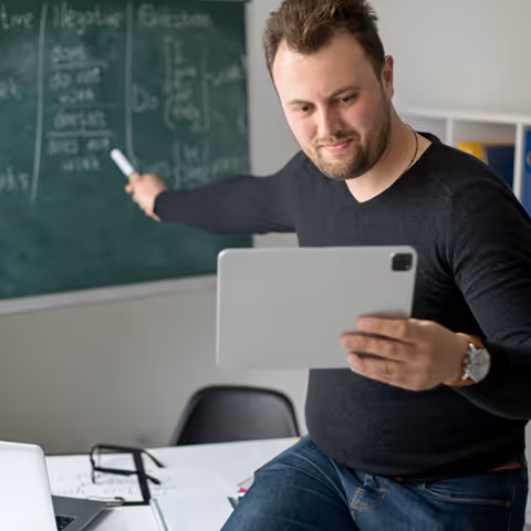 An online tutor sitting on a chair, holding a tablet, and pointing to a blackboard while tutoring students.