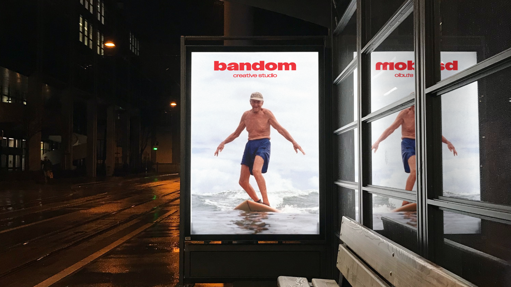 Backlit advertisement at night showing an elderly man surfing, with the text 'bandom creative studio' above him.