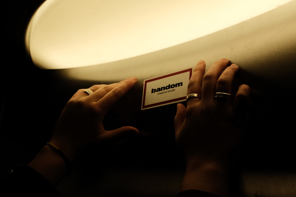 Two hands with rings holding a card that reads 'bandom creative studio' under soft warm light.