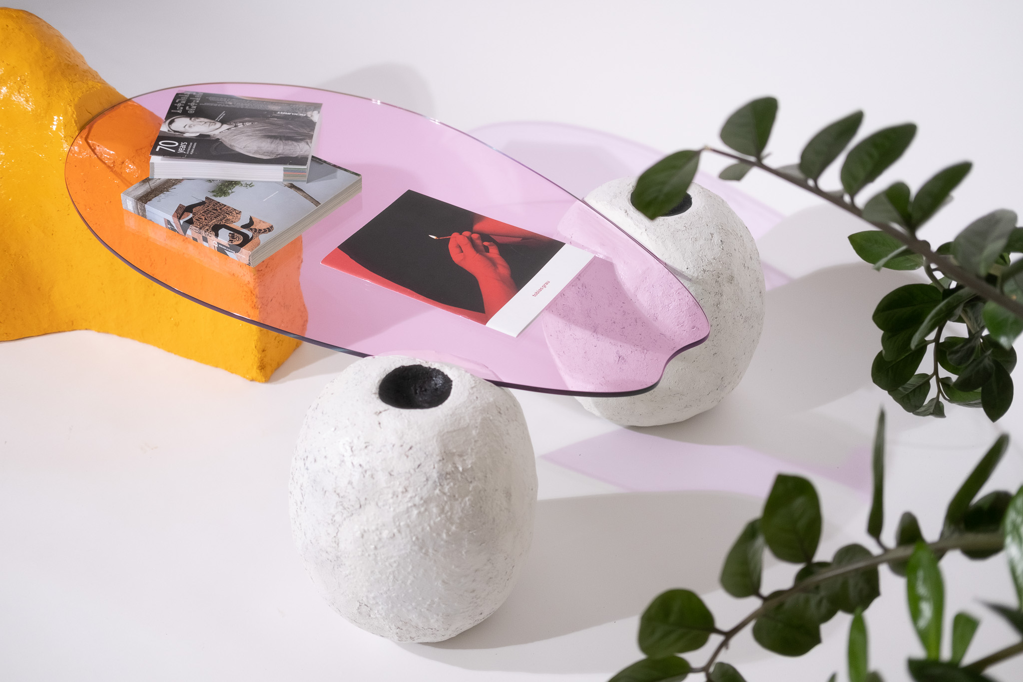 Modern purple glass tabletop supported by two textured white stone bases and a bright yellow support, with books on the table and green leaves in the foreground.