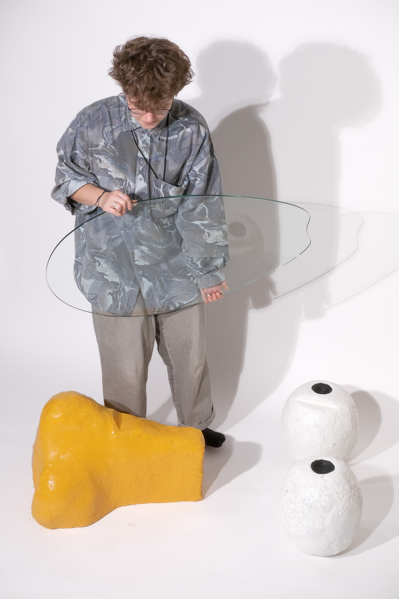 Person with curly hair and glasses holding an irregular-shaped glass sheet, standing near a large yellow abstract sculpture and two white oval sculptures with black circular tops.