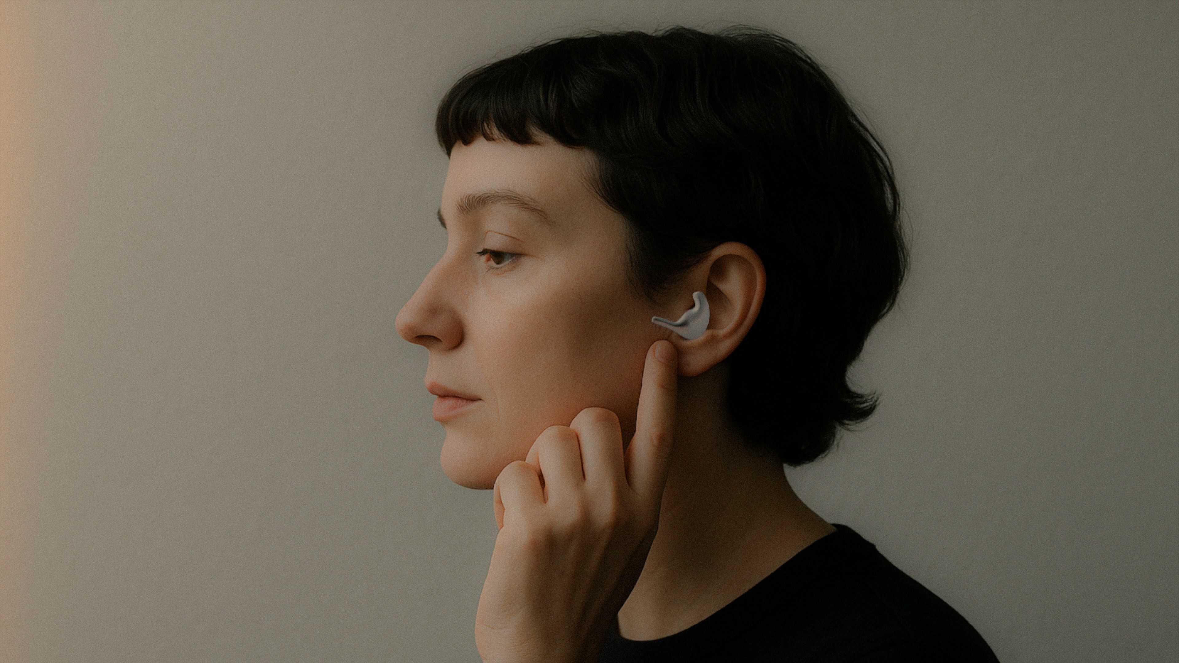 Side profile of a person adjusting a gray hearing aid in their ear against a plain background.
