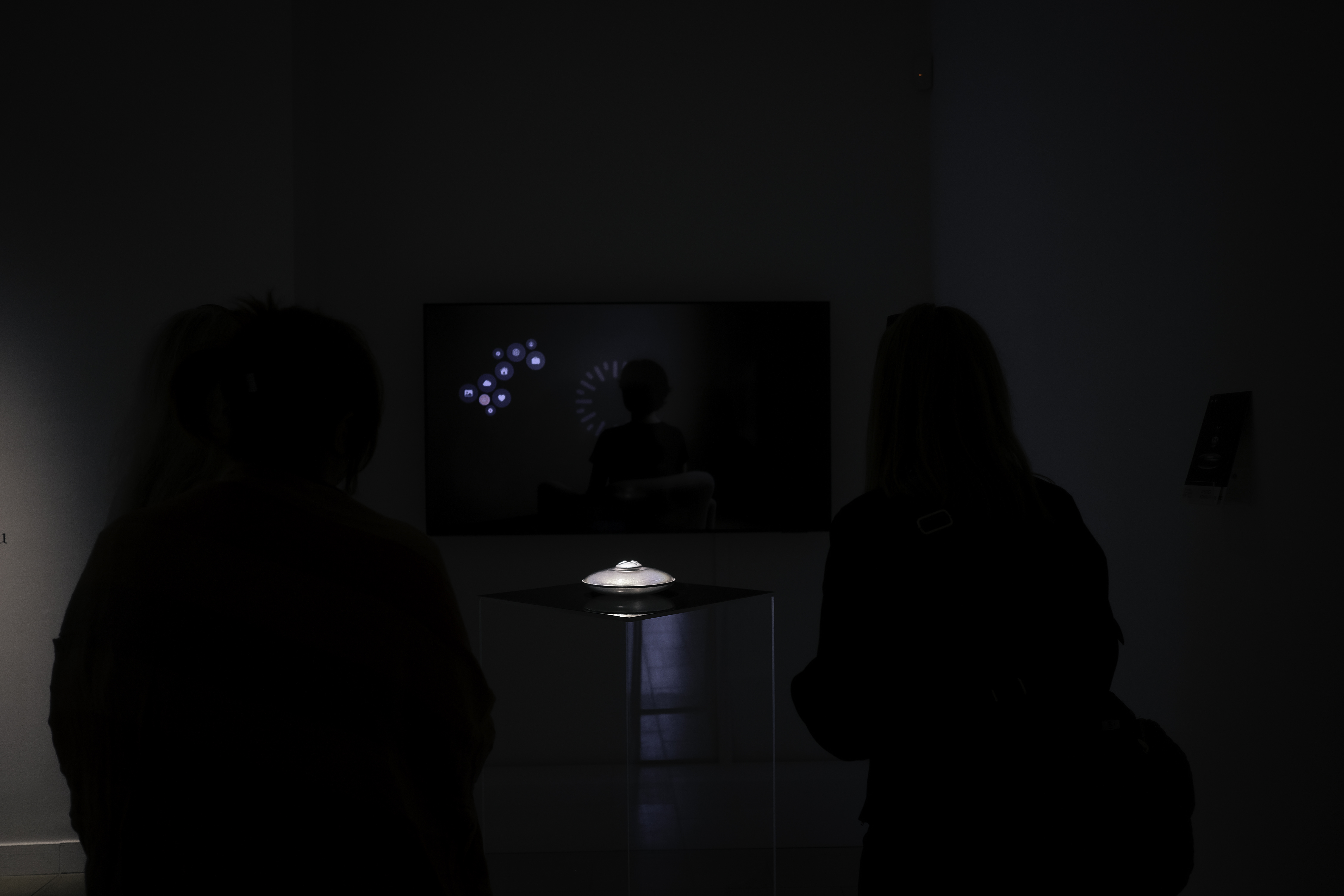 Silhouettes of three people viewing a small illuminated object displayed on a pedestal in a dark room with a screen showing a person and icons in the background.