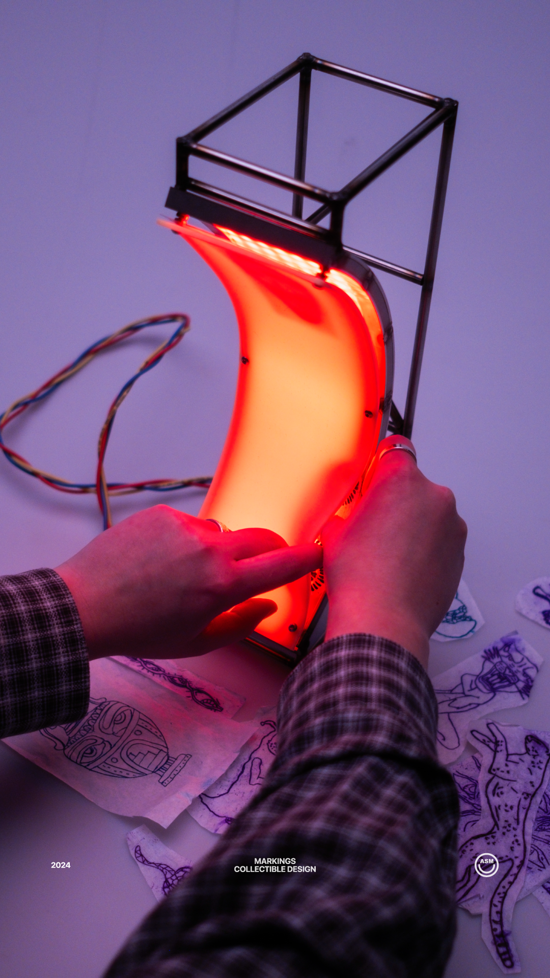 Person manipulating a flexible, glowing orange light panel mounted on a metal frame, surrounded by hand-drawn sketches on paper.