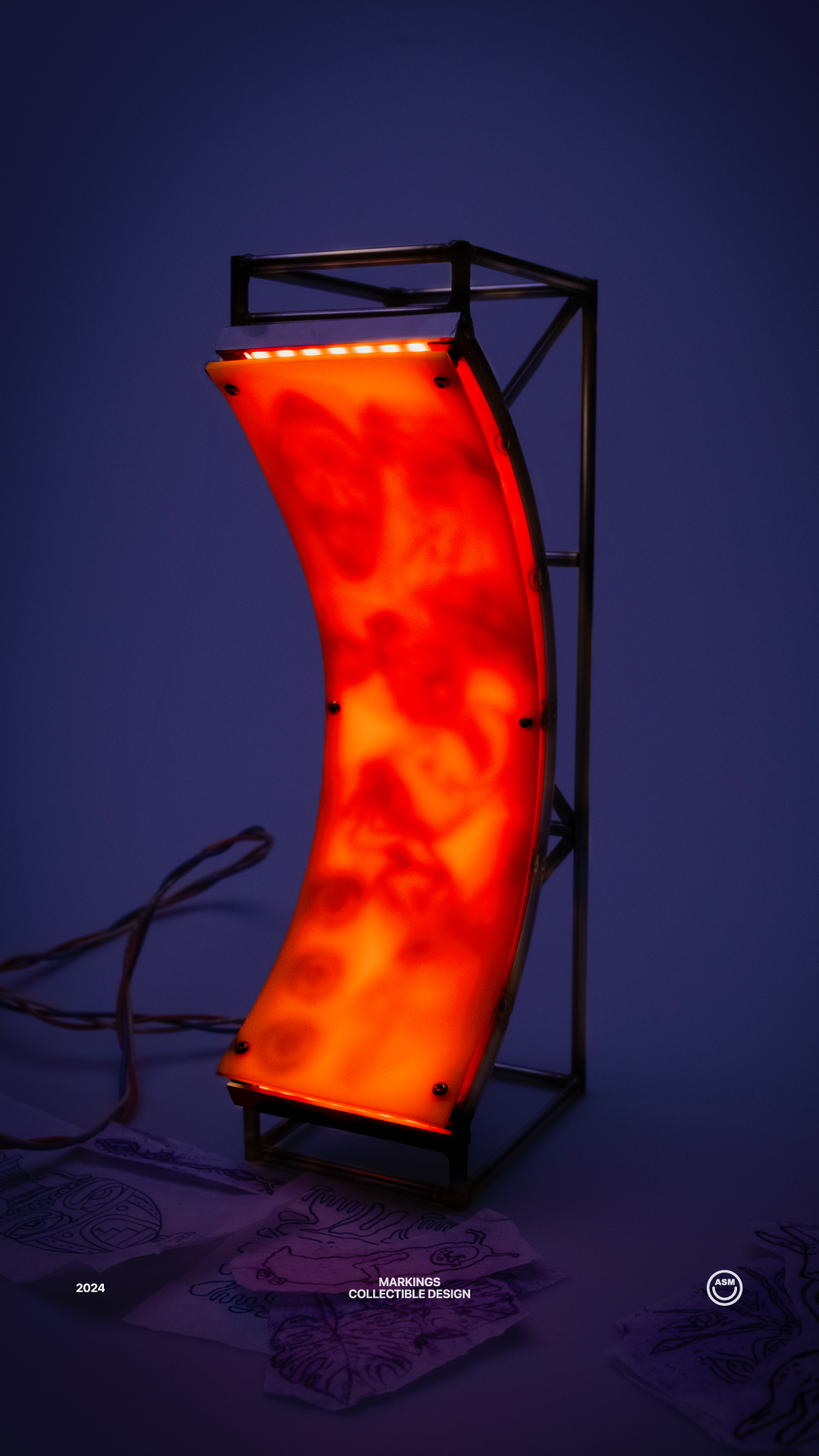 Curved orange illuminated panel with abstract shapes mounted on a metal frame, with sketches on paper beneath it against a dark blue background.