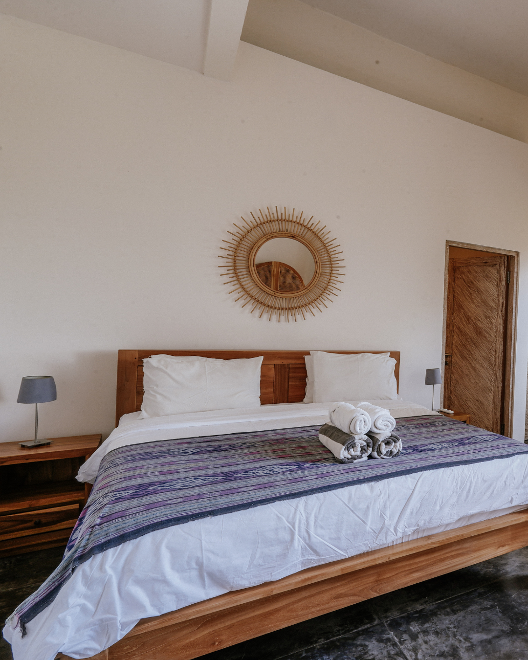 A neatly made wooden bed with white bedding and a purple patterned throw blanket, two white pillows, and four rolled towels stacked at the center, with a round rattan mirror above the headboard against a plain beige wall, flanked by two bedside tables each holding a small gray lamp, with a wooden door to the right.