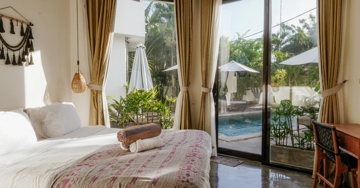 Bright bedroom with a bed covered in white sheets and a patterned bedspread, topped with two folded towels, next to large glass doors with beige curtains overlooking a lush garden and an outdoor pool with umbrellas and lounge chairs.