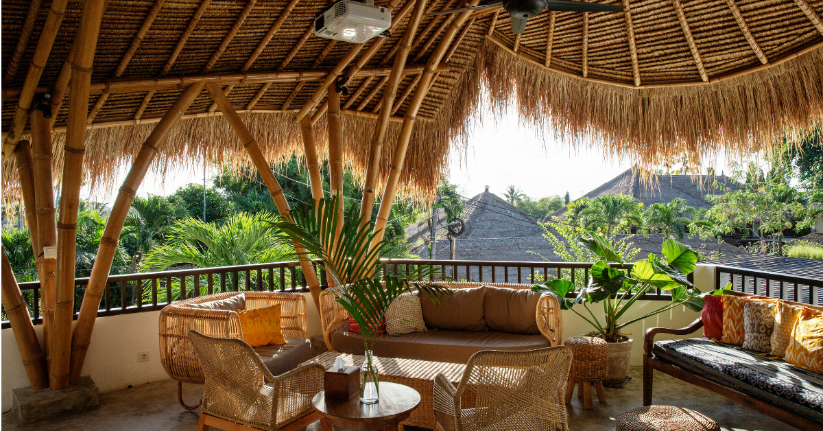 Outdoor seating area with bamboo furniture and wicker chairs under a thatched roof, decorated with green plants and colorful cushions, overlooking tropical trees and huts.