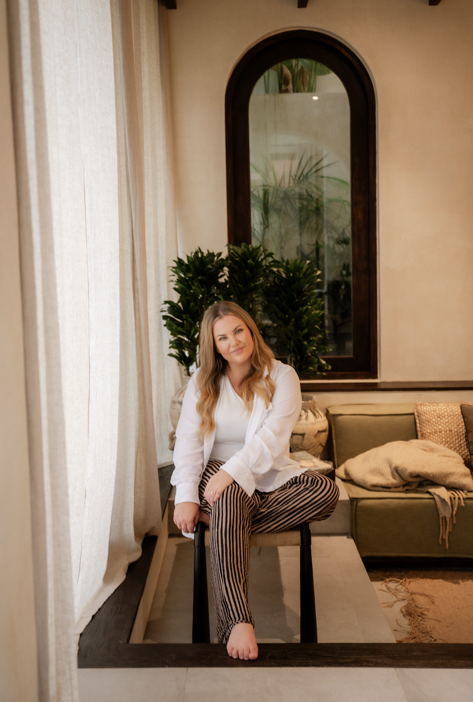 A woman with long blonde hair sitting barefoot on a chair in a cozy room with beige curtains, a green sofa with cushions and a blanket, and a large arched window with plants behind it.