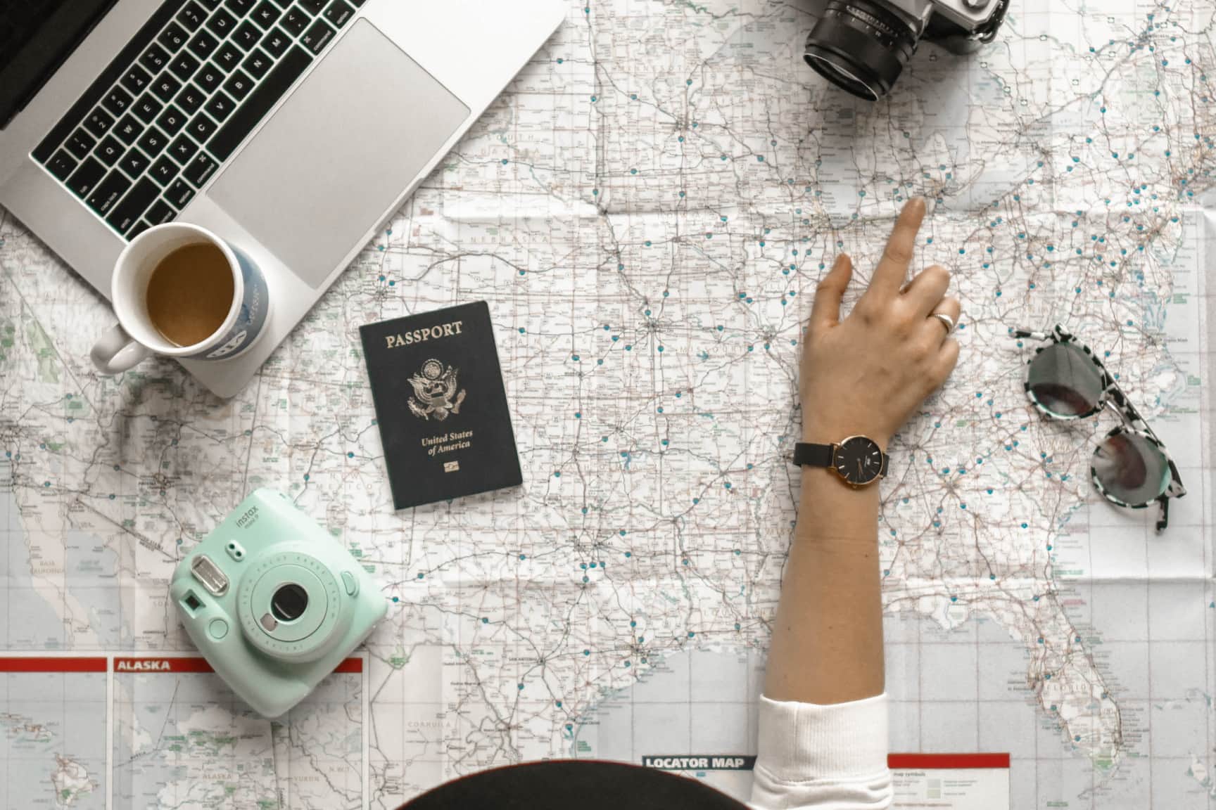 Overhead view of a hand pointing on a map of the United States surrounded by a laptop, coffee cup, passport, camera, and sunglasses.