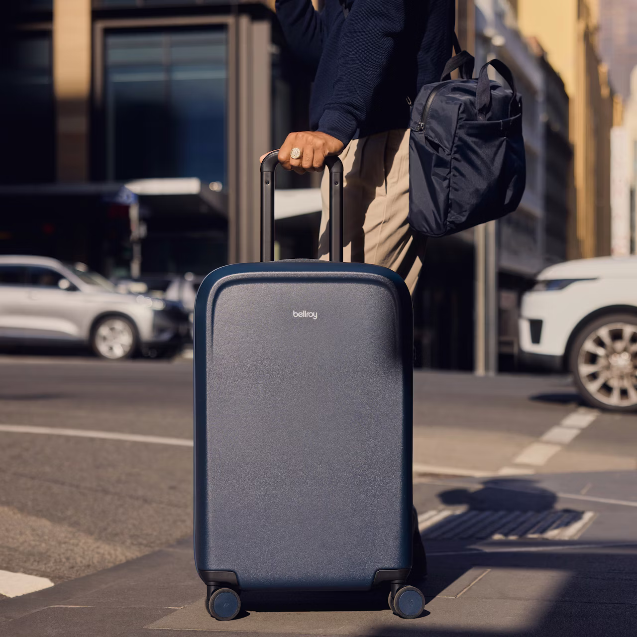 Person holding black Bellroy suitcase with extended handle and carrying a black bag walking on city street.