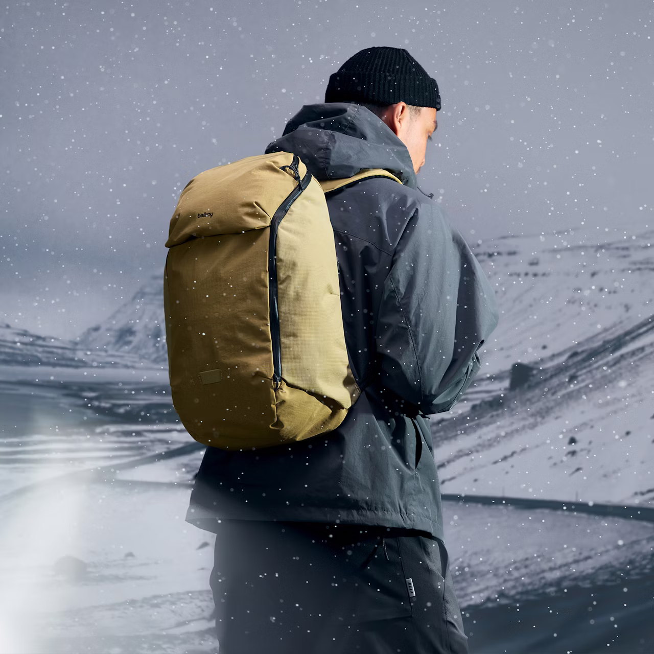 Person wearing a black beanie and dark jacket carrying a tan backpack in a snowy mountainous landscape.