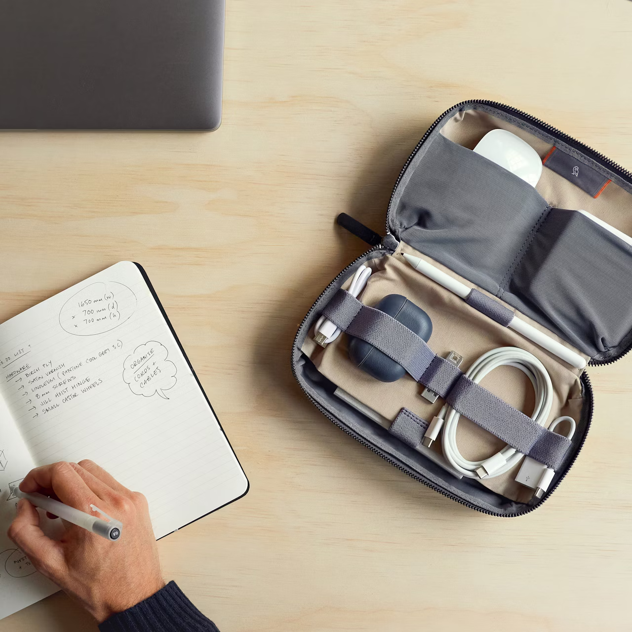 Open TechKit organizer pouch with cables, AirPods, and stylus on wooden desk next to a notebook and person's hand writing.