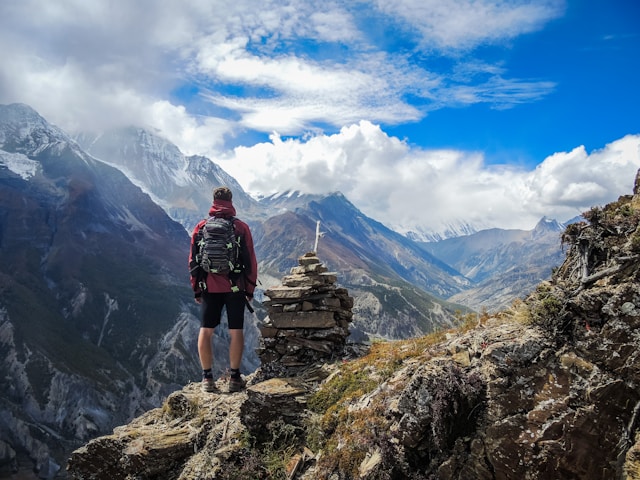 Hiker with backpack standing on rocky mountain edge overlooking a valley with snow-capped peaks under a partly cloudy blue sky.