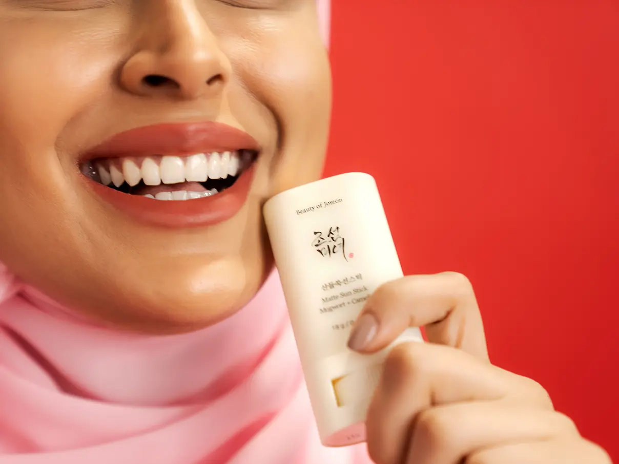 Closeup of a smiling woman's face. She is holding an iHerb product next to her face.